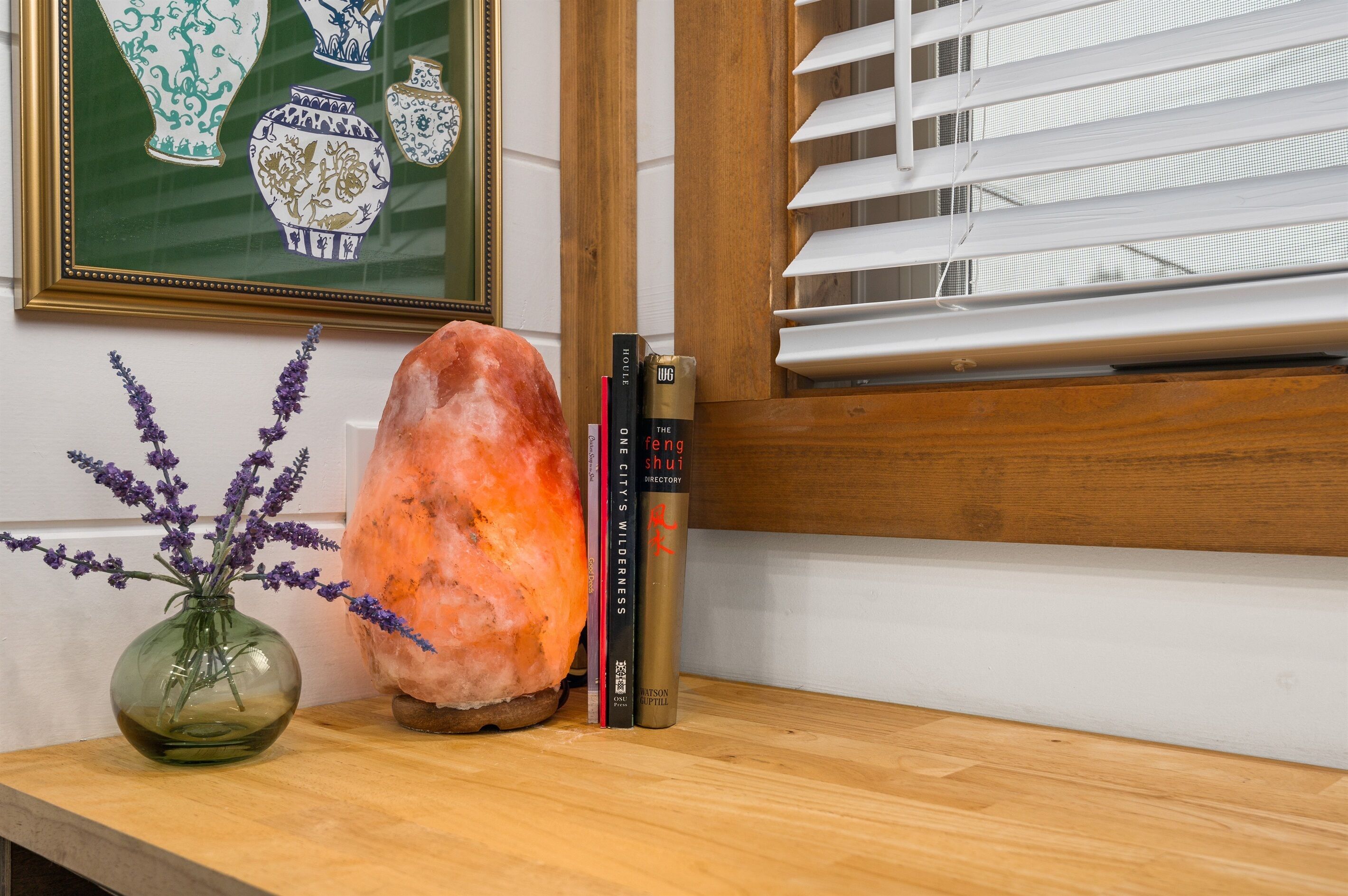 A wooden shelf with a Himalayan salt lamp, a glass vase with lavender, and a framed picture of vases.