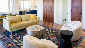 Sala de estar con un sofá, un sillón y una mesa de centro. Se observa una cocina al fondo con armarios blancos y una ventana.