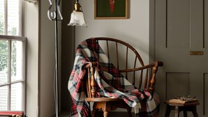 Een knus ingerichte kamer met een houten schommelstoel, een klein tafeltje met boeken en glazen, een lijst met een poster aan de muur en een raam met gordijnen.