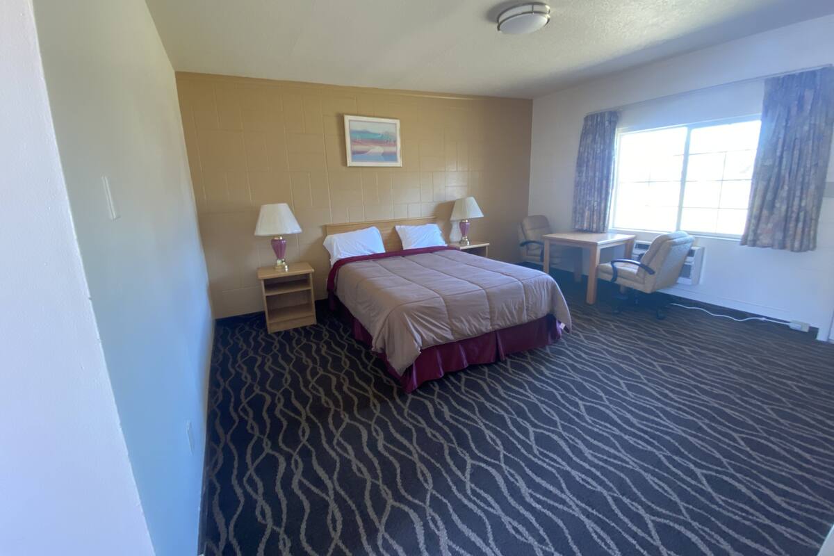 A hotel room with a bed, two bedside tables with lamps, a desk with a chair, a window with curtains, and a patterned carpet.