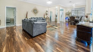 Amplia sala de estar con piso de madera, un sofá seccional gris y un comedor con candelabro.