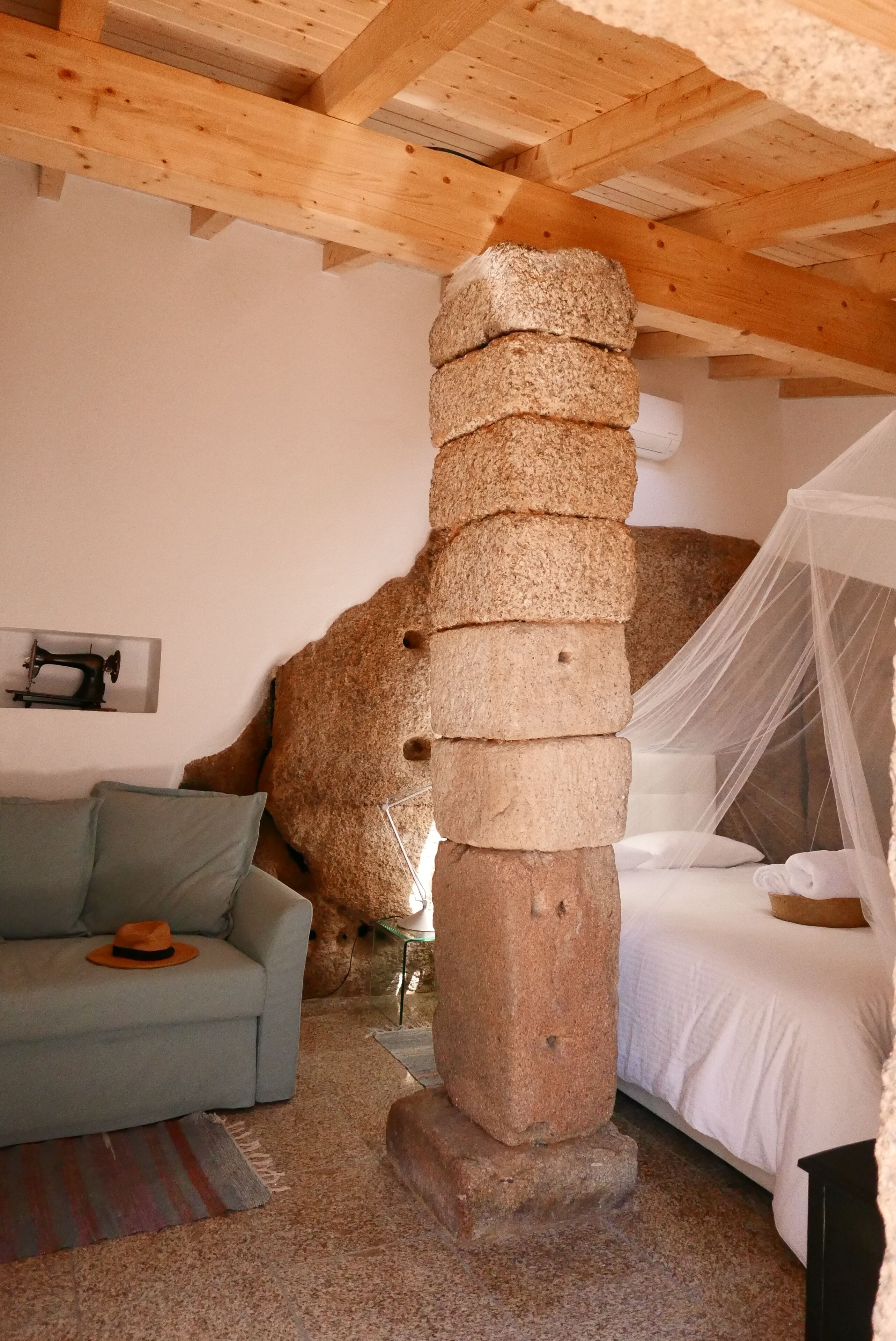 Quarto rústico com um pilar de pedra, uma cama com dossel, um sofá e uma máquina de costura na parede.