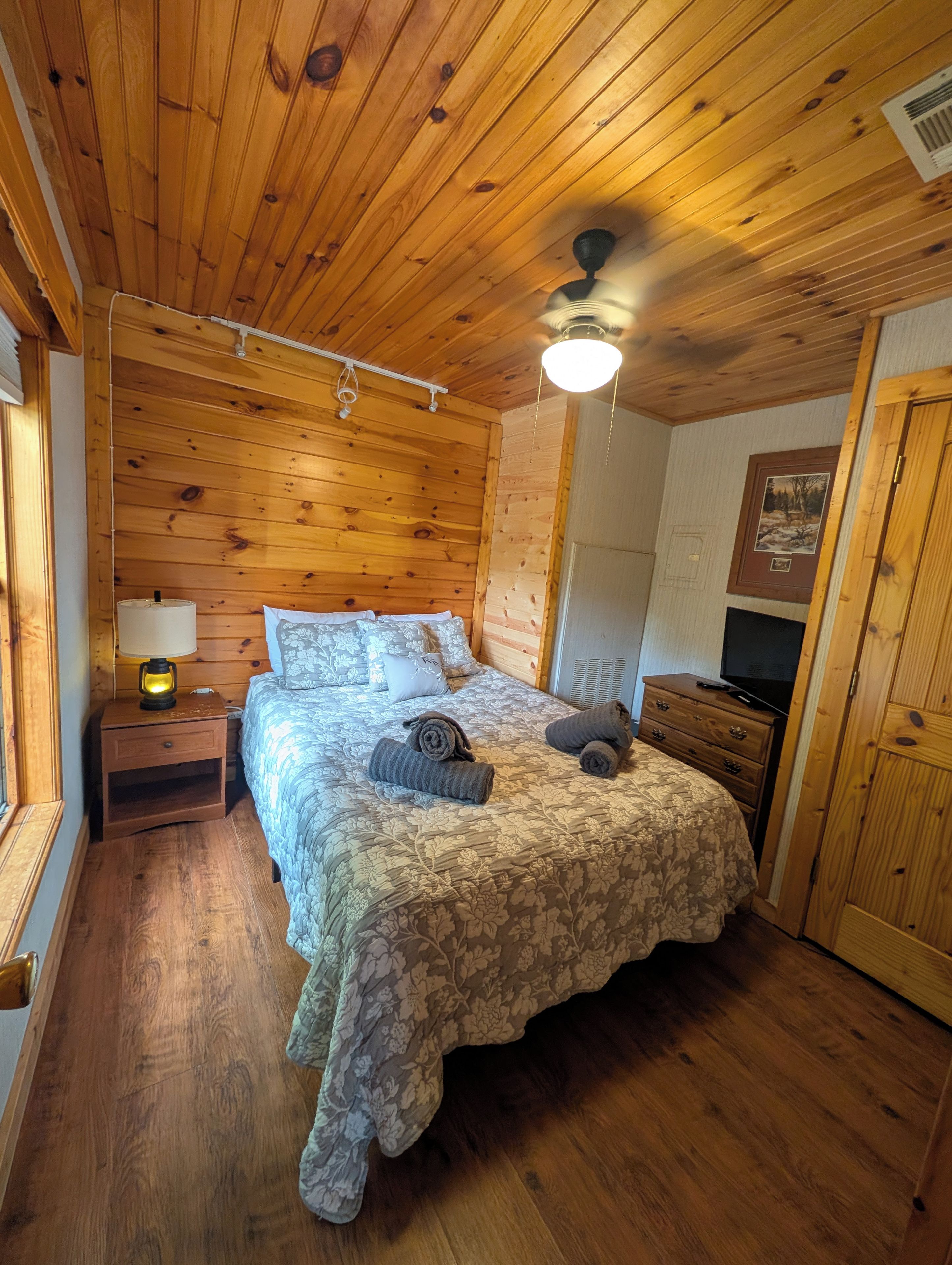 A wooden-paneled room with a bed, a nightstand with a lamp, a dresser, and a ceiling fan.