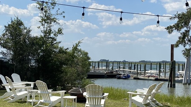 A grassy area with white Adirondack chairs and a string of lights against a backdrop of a clear sky and a marina with boats.