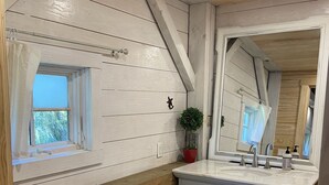 A bathroom with a wooden sloped ceiling, a window, a sink with a mirror, and a small potted plant.