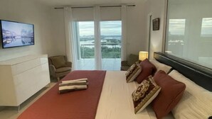 A modern bedroom with a large bed, a ceiling fan, a TV, and a view of a cityscape through large windows. - CONDO CORA. BLUE MARINE. SINT MAARTEN  (Sint Maarten)