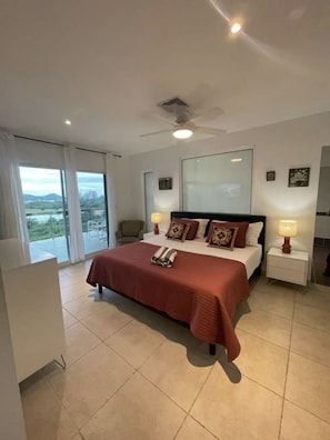 A bedroom with a large bed, two bedside tables with lamps, a ceiling fan, and a view of the outdoors through a sliding glass door.