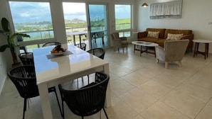 A spacious living area with a dining table and chairs, a sofa, and large windows offering a view of the outdoors. - CONDO CORA. BLUE MARINE. SINT MAARTEN  (Sint Maarten)