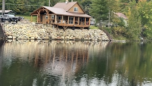 A wooden cabin with a deck, situated by a lake with a stone wall and a dock.