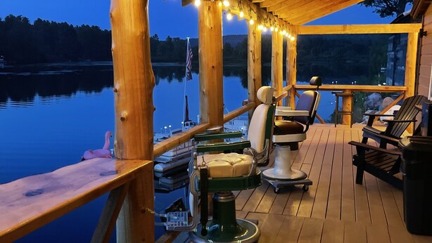 A wooden deck with outdoor seating, a coffee machine, and a view of a lake at dusk.