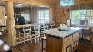 A wooden cabin interior with a kitchen area, dining space, and a large window offering a view of the outdoors.