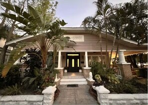 Uma casa com varanda frontal, cercada por plantas tropicais e palmeiras.