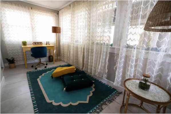 Quarto com escrivaninha, cadeira, dois almofadas, uma pequena mesa redonda com uma planta e cortinas de renda.
