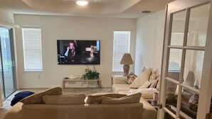 A living room with a large beige sectional sofa, a television mounted on the wall, and a door with glass panels. - The Villages
Lake Sumpter
Most desirable area 
Golf, pools and music every night (The Villages)
