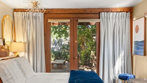 A bedroom with a bed, a wooden headboard, a nightstand, a lamp, a bench outside, and a view of greenery. - Adorable Santa Fe 3 Bedroom Home! (Santa Fe)