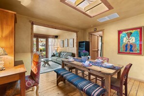 A cozy dining area with a wooden table, benches, and a colorful chair. There is a painting on the wall and a window with curtains. - Adorable Santa Fe 3 Bedroom Home! (Santa Fe)