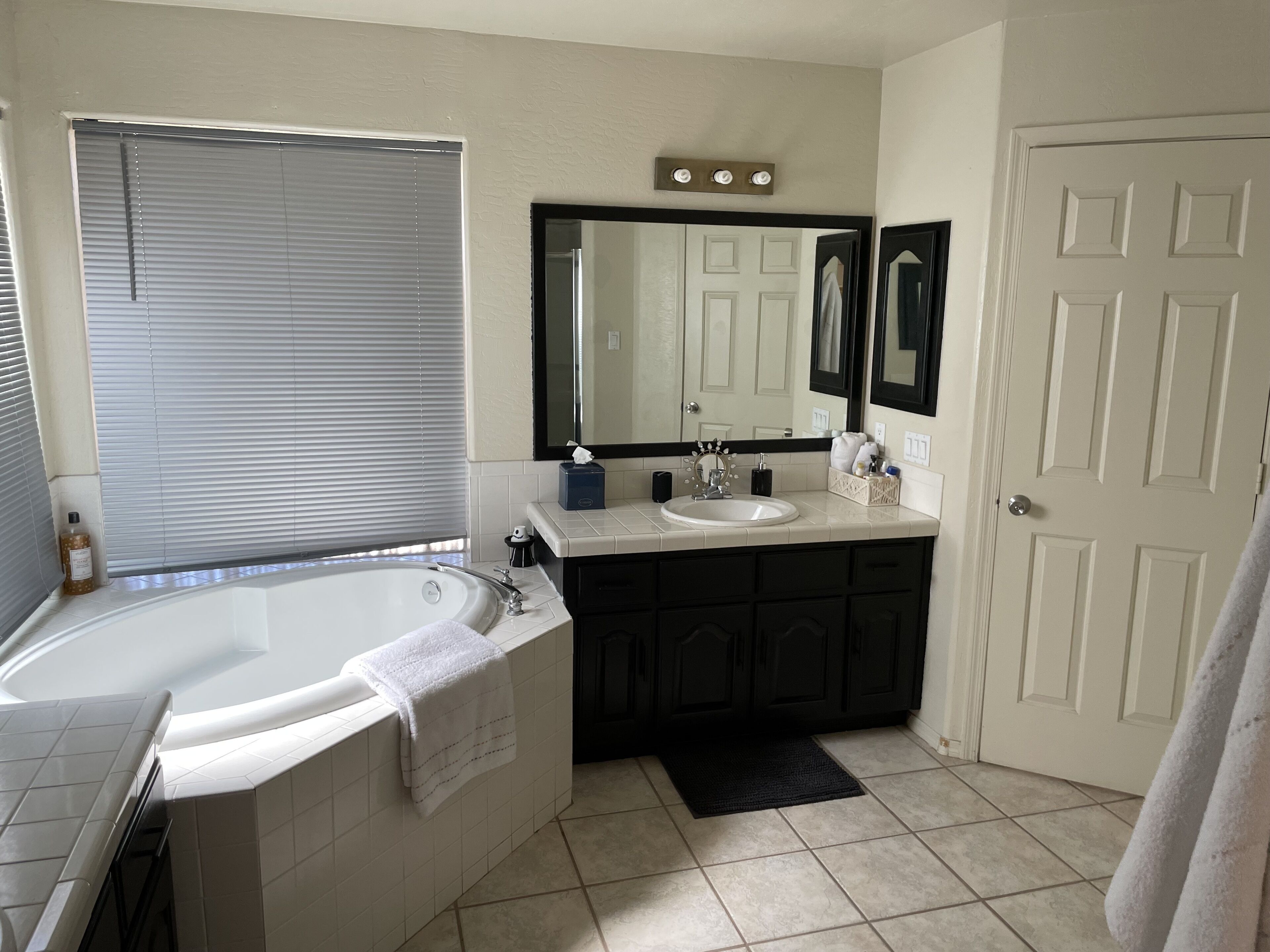 Une salle de bain avec une baignoire, un lavabo, un meuble-lavabo avec des placards sombres, un grand miroir et une porte.