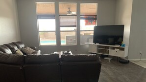 A living room with a leather sectional sofa, a flat-screen TV on a white entertainment unit, and a large window with blinds. - Backyard Escape with no cleaning fee or extra guest fees (Yuma)