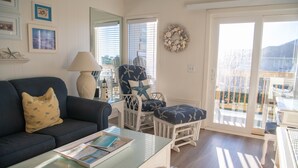 Un salon avec une table basse en verre, un canapé bleu foncé, un fauteuil blanc orné d’un motif en forme d’étoile de mer, et une vue sur l’extérieur grâce à une porte-fenêtre coulissante.
