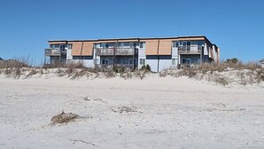 Un immeuble d’appartements situé en bord de mer, doté de nombreux balcons, avec une plage de sable au premier plan.