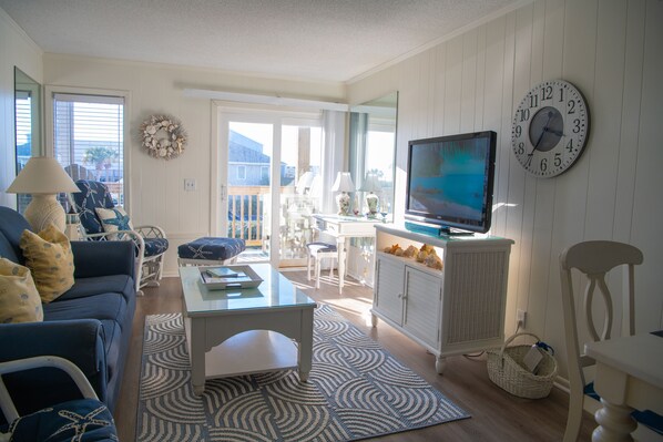 A living room with a sofa, coffee table, TV, and a view to the outside. - Ocean View, 2 Bedroom, 2 Bath with direct beach access and pool (Ocean Isle Beach)