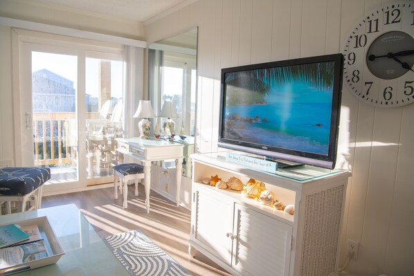 A living room with a television, white furniture, a glass coffee table, and a view of the outdoors. - Ocean View, 2 Bedroom, 2 Bath with direct beach access and pool (Ocean Isle Beach)