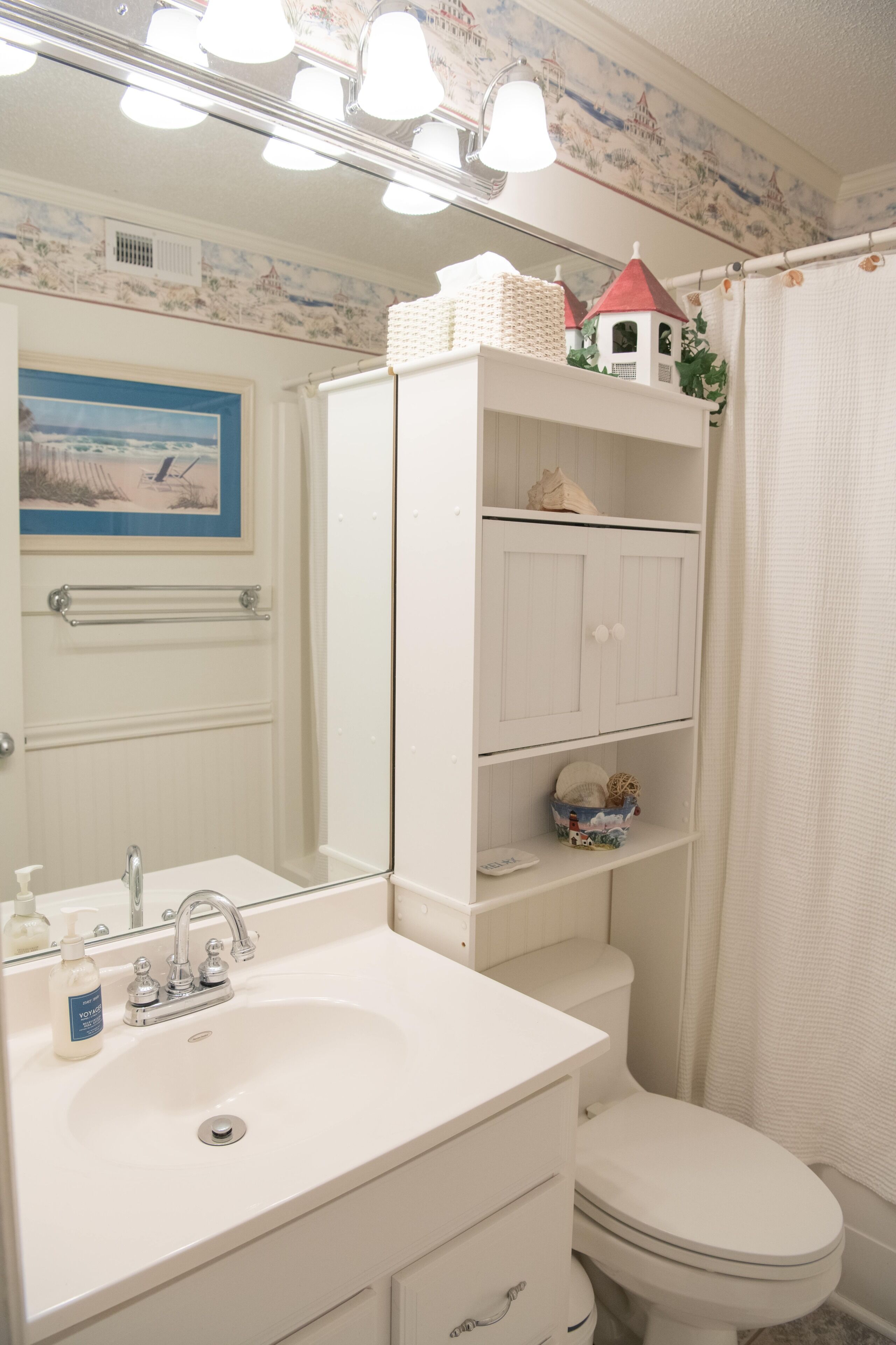 Casa de banho com lavatório branco, espelho e sanita. Existem prateleiras sobre a sanita com objetos decorativos.