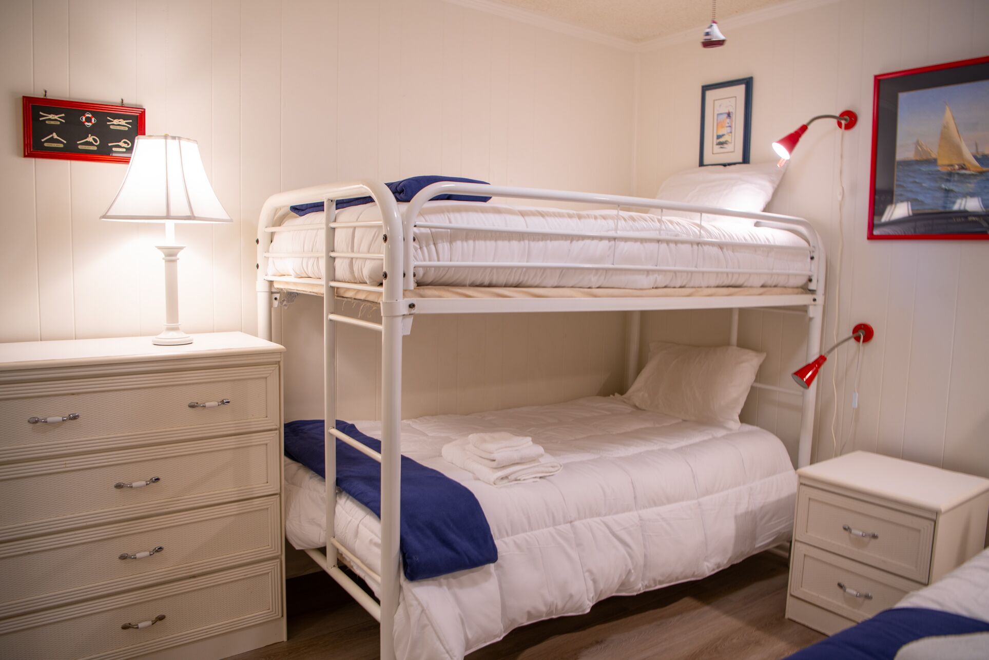 A bunk bed with a bedside table and a lamp, a dresser with drawers, and a framed picture on the wall.