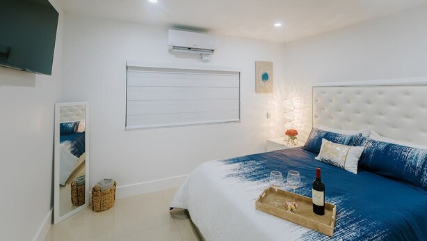 A bedroom with a large bed, a television mounted on the wall, an air conditioning unit, and a bedside table with a bottle and glasses. - Adorable Studio (Miami)