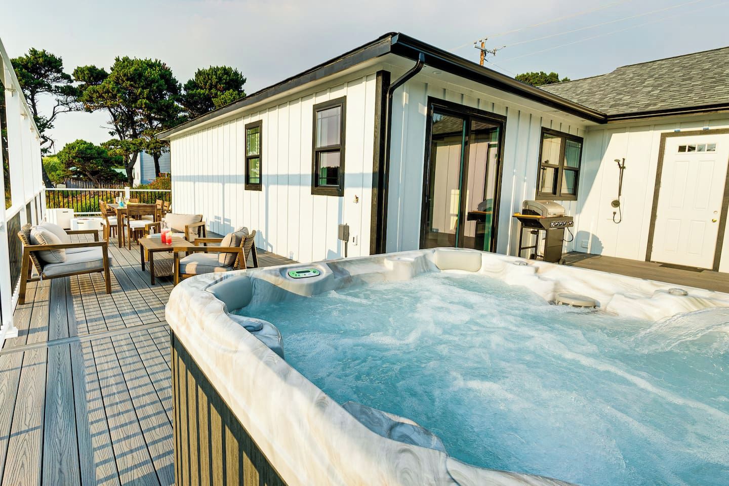 A hot tub with a wooden deck and outdoor furniture, a white building with windows and a door, and a fenced yard with trees.