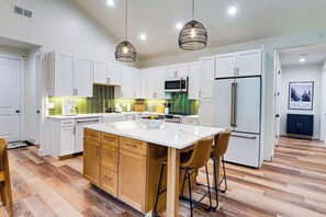 A modern kitchen with white cabinets, a central island, wooden bar stools, and pendant lighting. - The Lily Pond House-Walk to Beach, SPA, Sleeps 6 (Gold Beach)