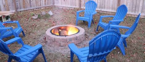 A backyard with a fire pit surrounded by blue plastic chairs.