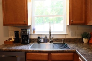 A kitchen with wooden cabinets, a stainless steel sink, a coffee maker, and a window with a view of greenery. - 3BR Pet-Friendly Retreat near Camp Lejeune (Jacksonville)