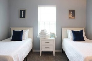 A room with two beds, each with a white headboard and a dark blue pillow. There is a small white nightstand with a drawer between the beds, and a window with blinds in the center.