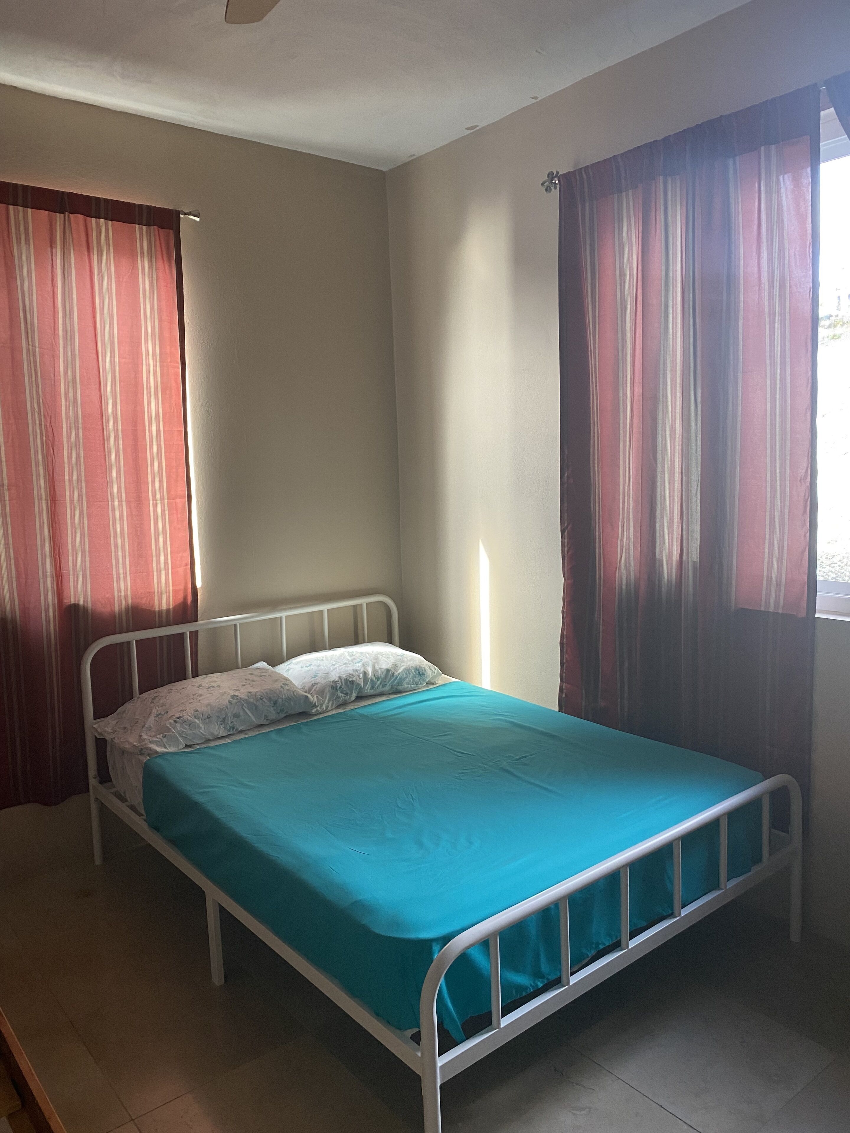 A single-bed room with a metal frame, a teal bedspread, and white pillows. There are two windows with striped curtains, one on each side of the bed.