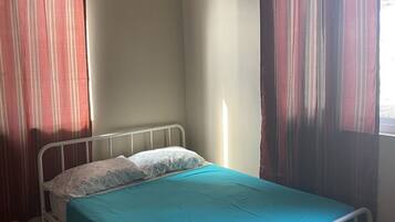 A single-bed room with a metal frame, a teal bedspread, and white pillows. There are two windows with striped curtains, one on each side of the bed.