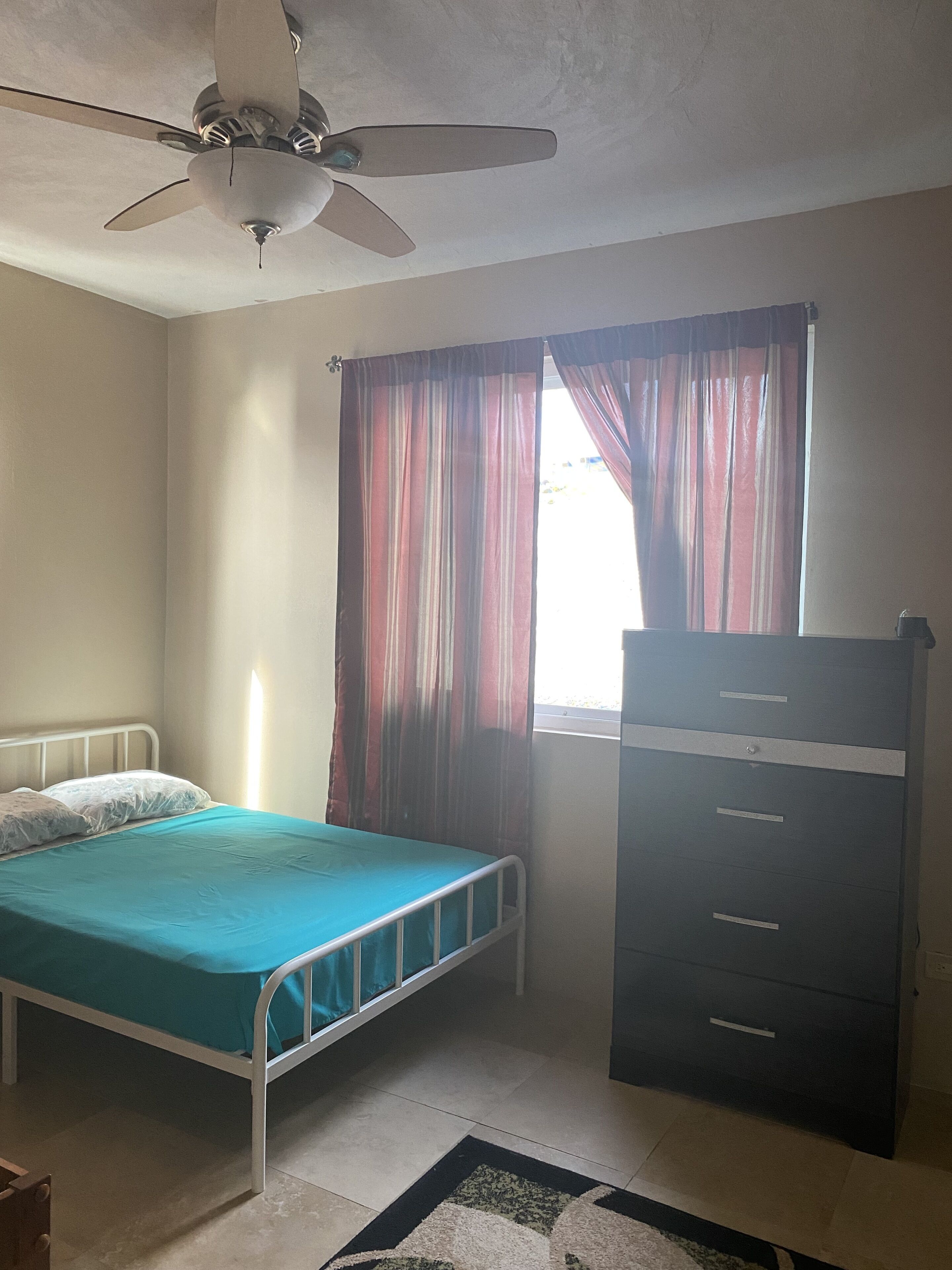 Een slaapkamer met een bed, een kapstok, een plafondventilator en een raam met gordijnen.