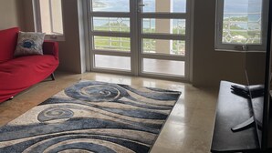 Un salon avec un canapé rouge, un tapis à motifs, un bureau avec un clavier et de grandes fenêtres donnant sur un paysage.
