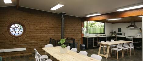 Une pièce spacieuse avec une grande table en bois et des chaises blanches, un mur en briques agrémenté d’une fenêtre circulaire, et un coin cuisine équipé d’appareils modernes.