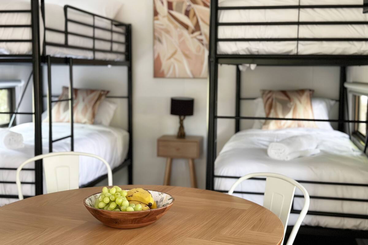 Um quarto com beliches, uma pequena mesa com uma tigela de frutas e uma luminária em uma mesa de cabeceira.