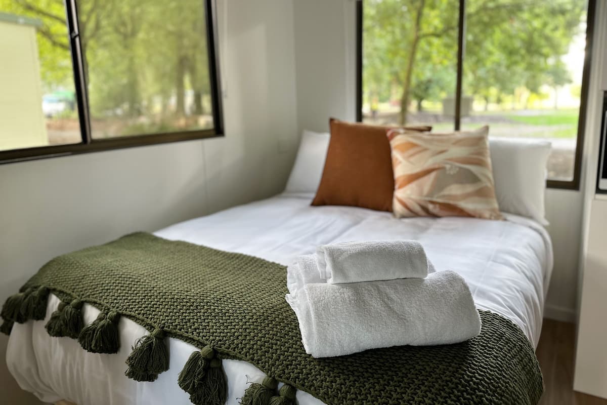 Cama bem arrumada com manta verde e travesseiros brancos, toalha branca sobre a cama, e vista para árvores através de grandes janelas.