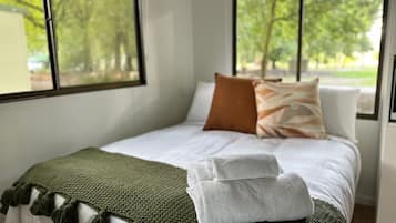 Un lit bien fait, recouvert d’une couverture verte et agrémenté d’oreillers blancs, une serviette blanche posée sur le lit, et une vue sur les arbres à travers de grandes fenêtres.