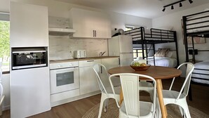 A compact kitchen with white cabinets, a microwave, a toaster, and a sink. There is a wooden table with chairs and a bowl of fruit. A bunk bed is visible in the background. - Tawonga Tourist Park (Tawonga)