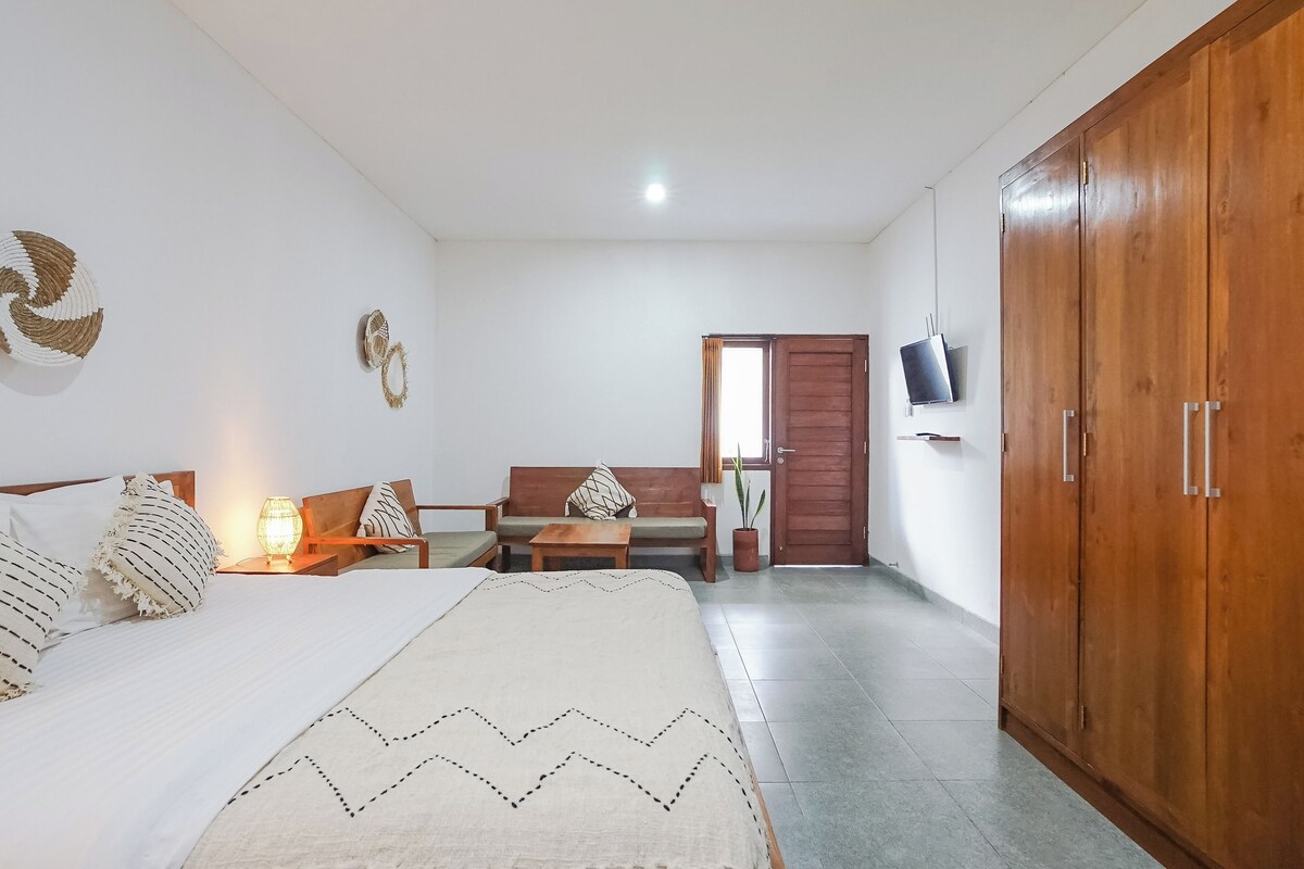 A bedroom with a bed, a wooden wardrobe, a sofa, a small table, and a TV mounted on the wall.