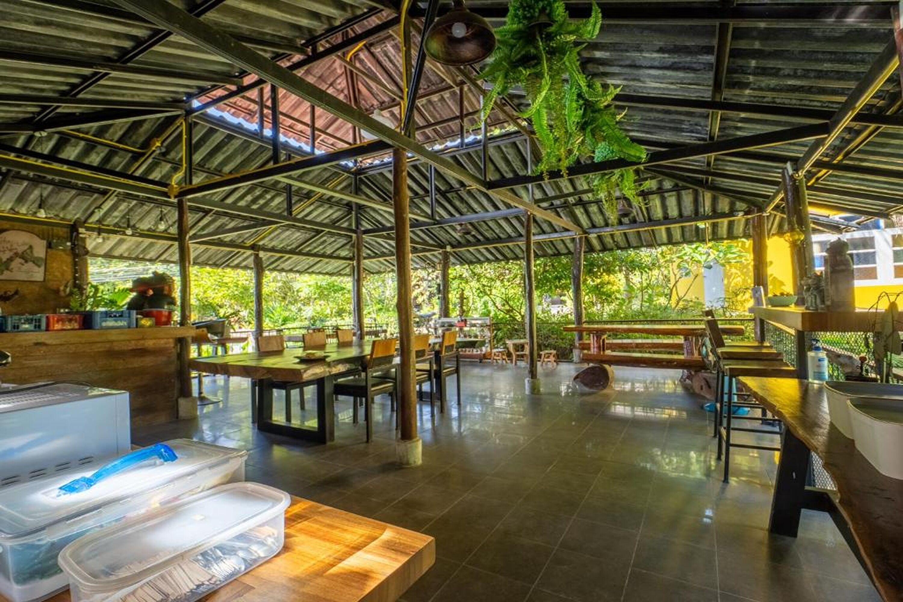 An open-air restaurant with wooden tables and chairs, a metal roof, and potted plants.