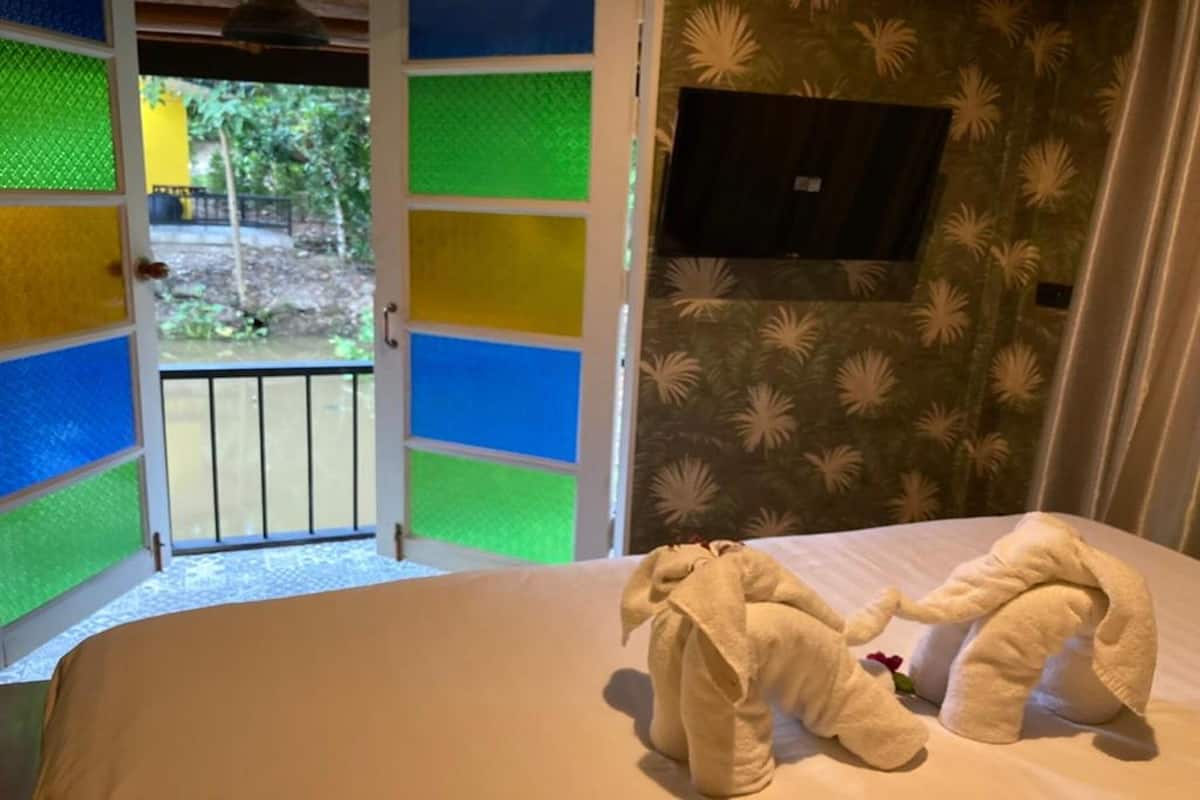 A room with a bed, two towels shaped like elephants, a window with a view of greenery, and a patterned curtain.