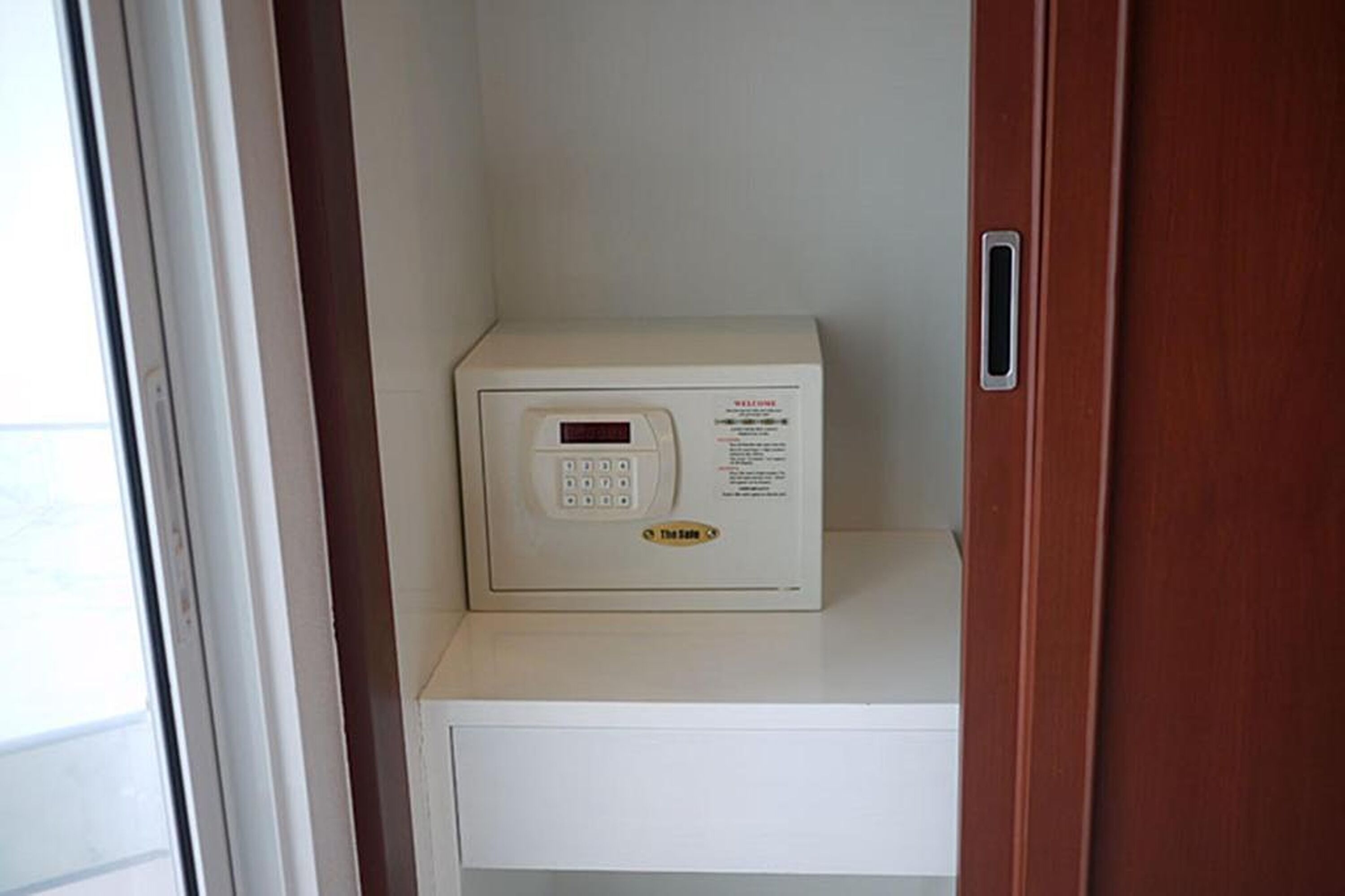 A white security keypad with a digital display, located on a white shelf in a room with a wooden door.
