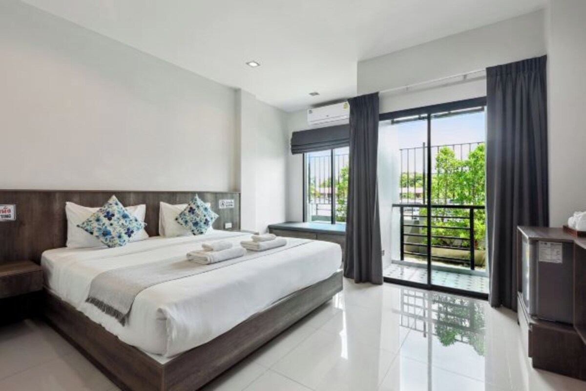 A hotel room with a large bed, a small desk, a refrigerator, and a balcony with a view of greenery.