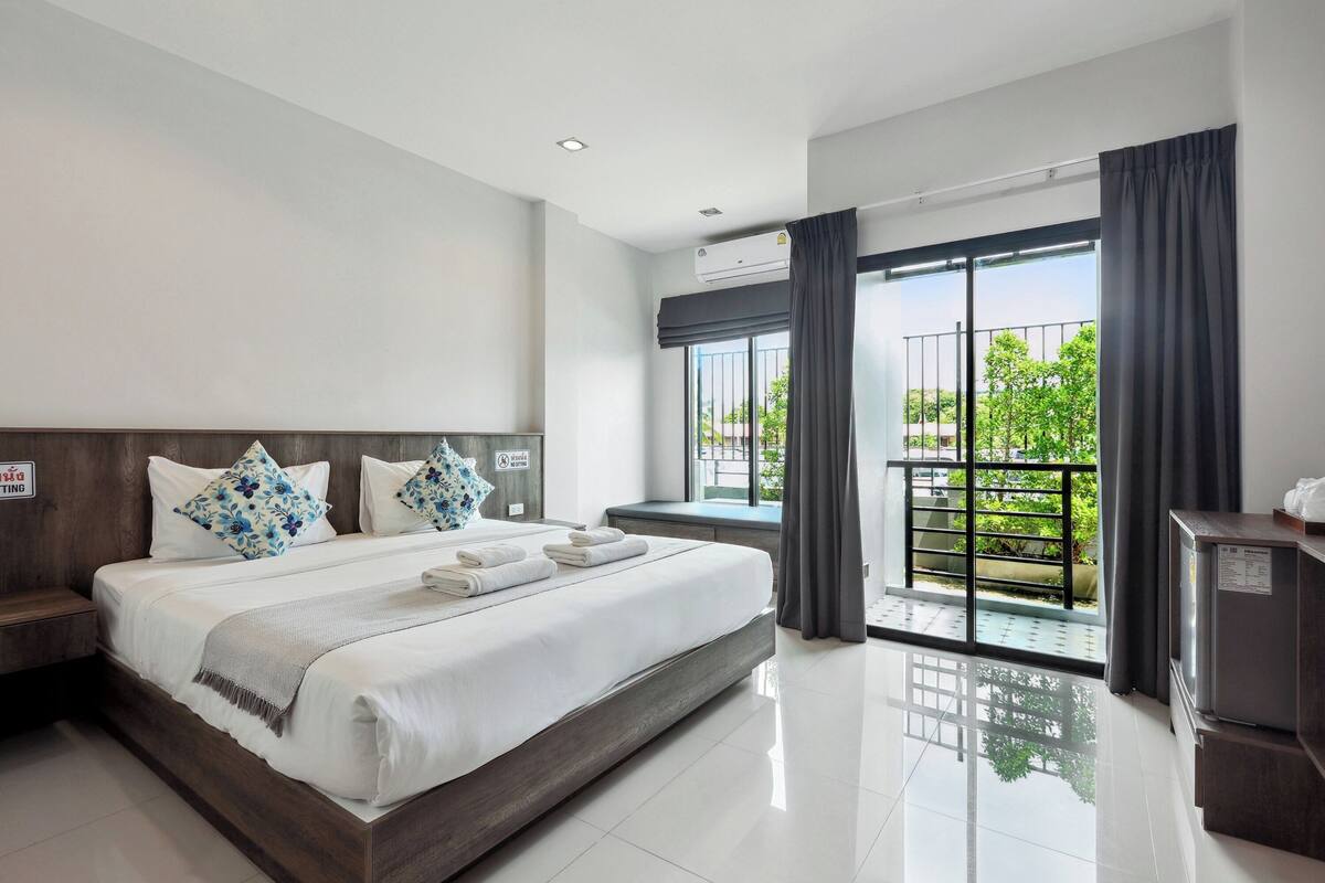 Een moderne hotelkamer met een groot bed, een klein bureau, een koelkast en een balkon met uitzicht op groen.