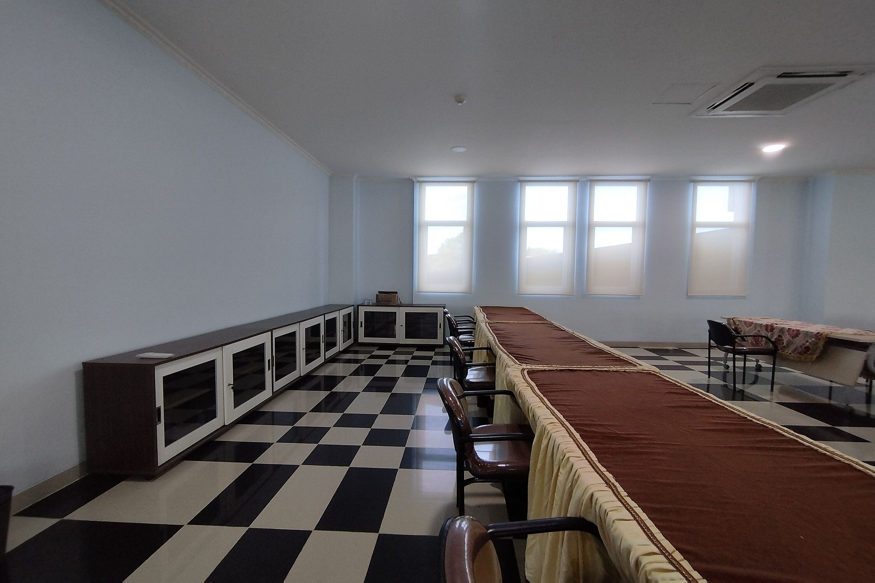 Una habitación con piso a cuadros, una mesa larga con sillas, un armario y un ventanal con persianas.